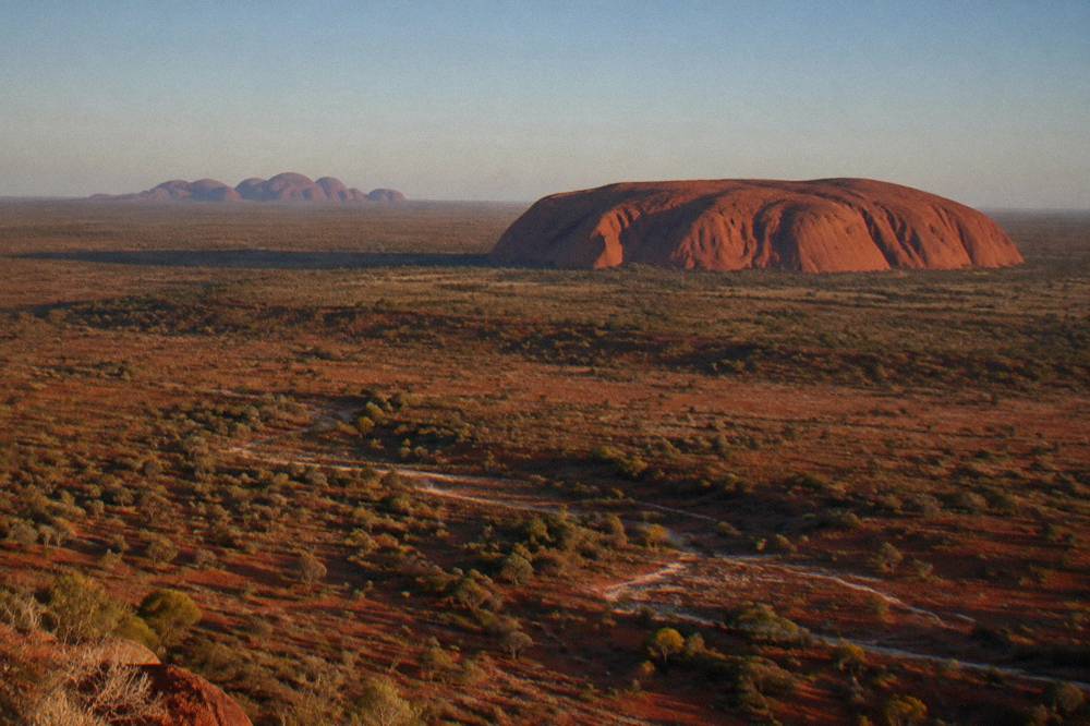Aerial view across red desert toward Uluru and distant domes of Kata Tjuta